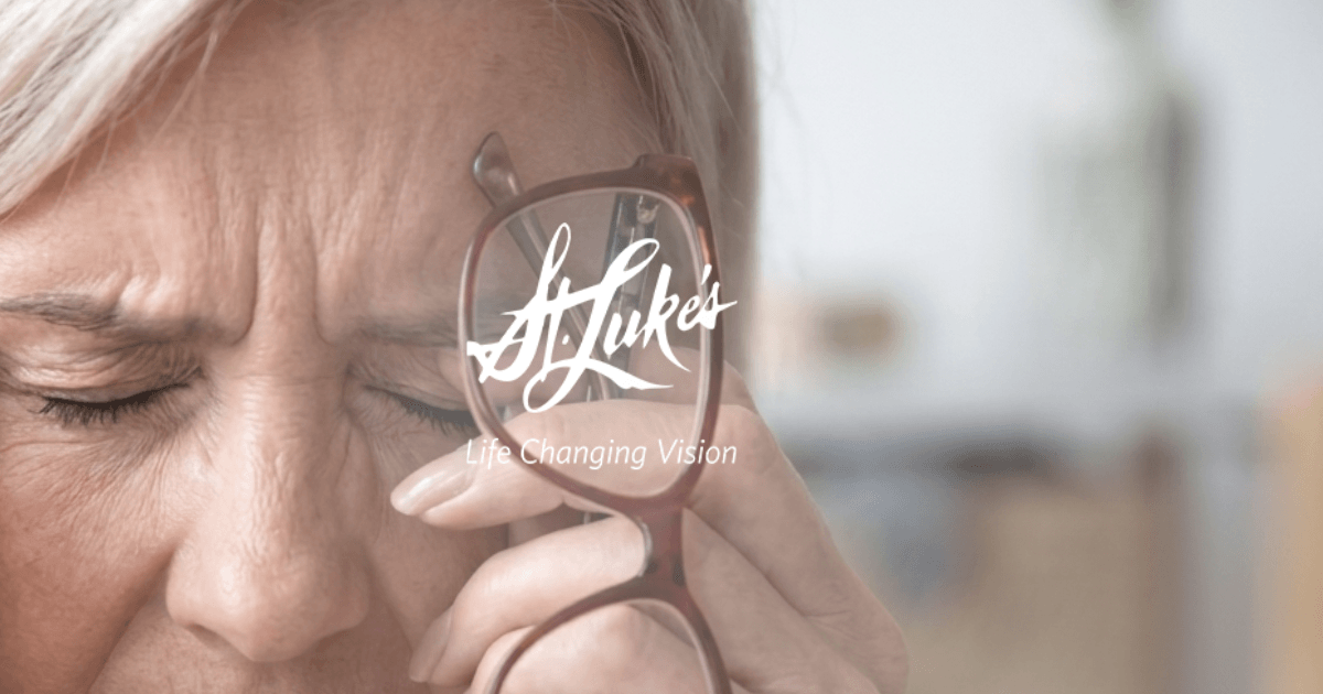 A close-up of an older woman, her face pained, eyes closed. She rubs her left temple with one hand and holds glasses in the other, contemplating cataract surgery.